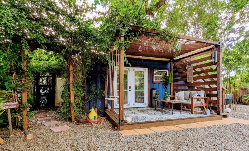 A cozy blue container home surrounded by greenery, featuring a wooden deck and outdoor seating area.