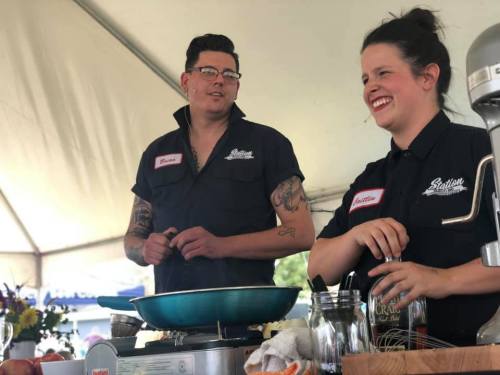 Two chefs smile and interact while cooking at a demonstration, with a skillet and ingredients on the table in front of them.