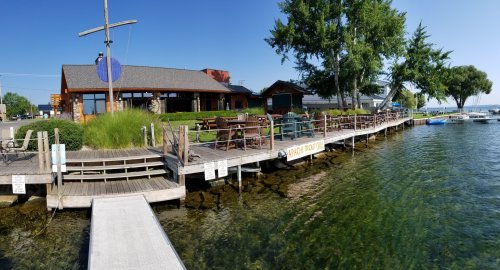 A wooden dock leads to a lakeside restaurant with outdoor seating, surrounded by greenery and clear blue water.