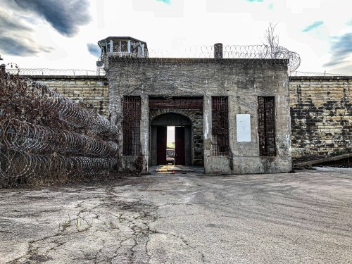 An old, abandoned prison entrance with barbed wire and a guard tower under a cloudy sky.