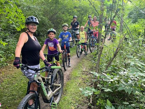 A group of children and adults on mountain bikes, smiling and posing on a trail surrounded by greenery.