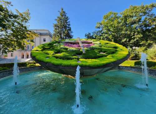 A large, circular floral clock surrounded by water fountains and greenery, set against a clear blue sky.