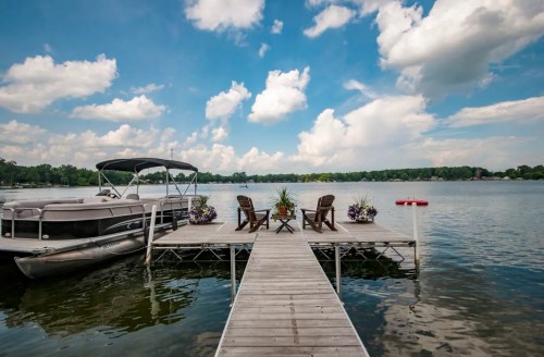 A wooden dock extends over calm water, featuring two chairs and potted plants, with a boat nearby and a blue sky above.