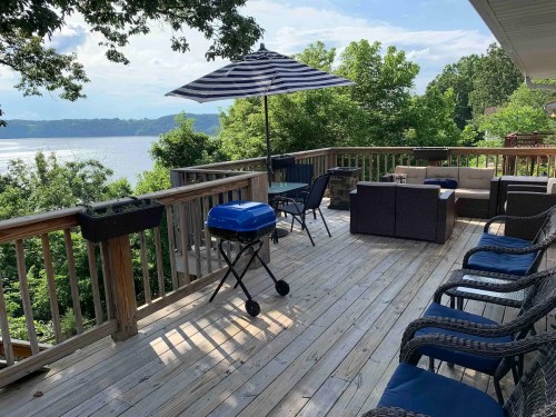 A spacious deck with a grill, seating area, and an umbrella, overlooking a scenic river and lush greenery.