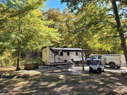 A camper and truck parked under trees in a serene outdoor setting, with a picnic table and fire pit nearby.