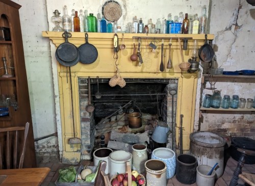 A rustic fireplace with colorful bottles, cooking utensils, and various pots arranged on a wooden mantel and floor.