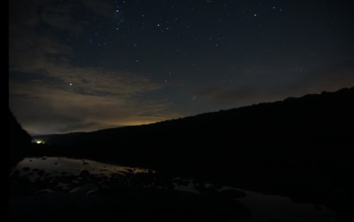 A serene night scene with a starry sky reflected in a calm river, surrounded by dark hills and soft clouds.