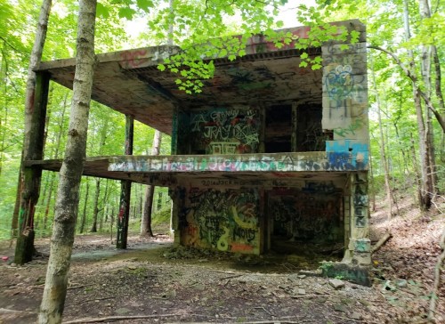 Abandoned two-story structure in a forest, covered in colorful graffiti and surrounded by lush green trees.