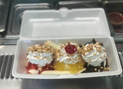 A takeout container with three scoops of ice cream topped with whipped cream, nuts, and a cherry, alongside fruit toppings.