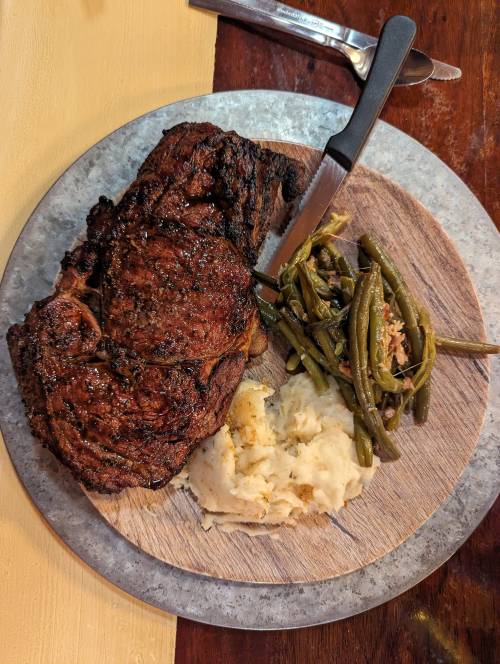 A plate with a large piece of grilled meat, mashed potatoes, and green beans, served on a wooden board.