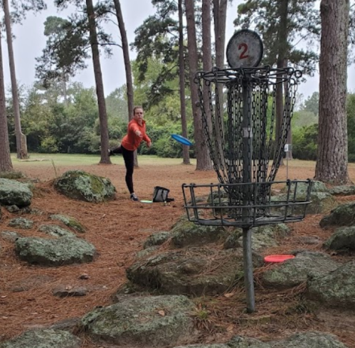 A person in an orange shirt throws a disc towards a metal disc golf basket in a wooded park setting.