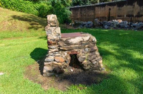 A rustic stone chair sits on green grass, surrounded by trees and a rocky backdrop.