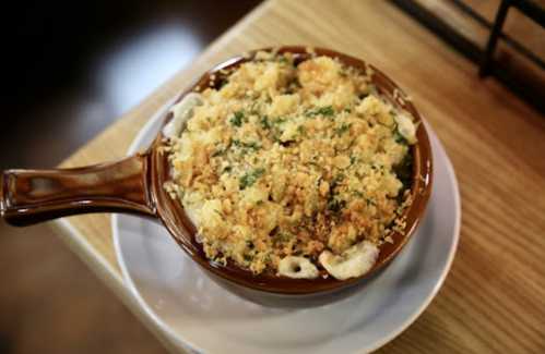 A baked dish of macaroni and cheese topped with golden breadcrumbs, served in a brown ceramic bowl on a white plate.