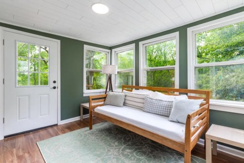 Bright sunroom with a wooden daybed, lamp, and large windows overlooking greenery. Light green walls and wooden flooring.