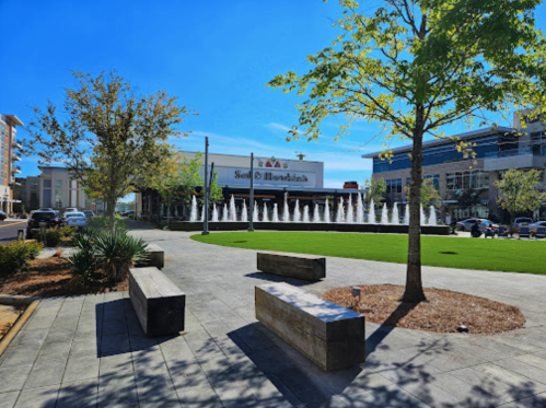 A sunny plaza with green grass, a fountain, and benches, surrounded by modern buildings and trees.