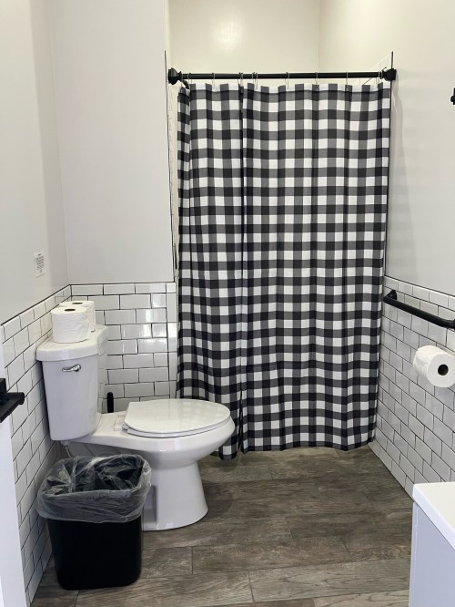 A clean bathroom featuring a black and white checkered shower curtain, toilet, trash can, and tiled walls.
