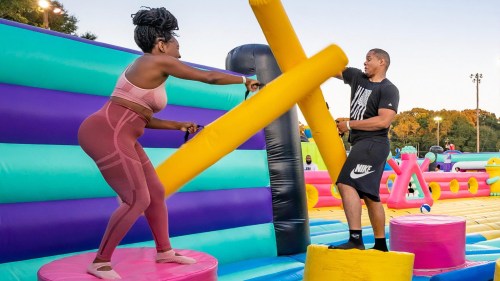 Two people engage in a playful competition on an inflatable obstacle course, using large foam poles.