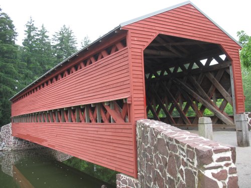 A red covered bridge with a wooden frame spans over a small body of water, surrounded by green trees.