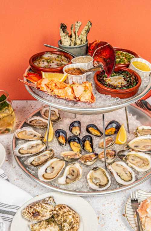 A tiered seafood platter featuring oysters, mussels, lobster, and various sauces, set against a vibrant orange background.