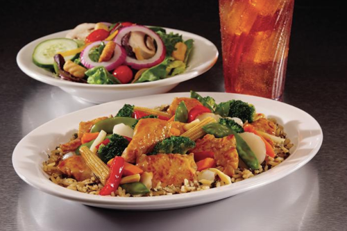 A plate of stir-fried chicken with vegetables over rice, alongside a fresh salad and a glass of iced tea.