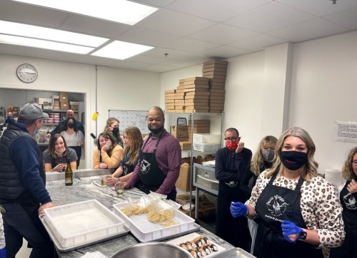 A group of people in an industrial kitchen, wearing aprons and gloves, preparing food and smiling at the camera.