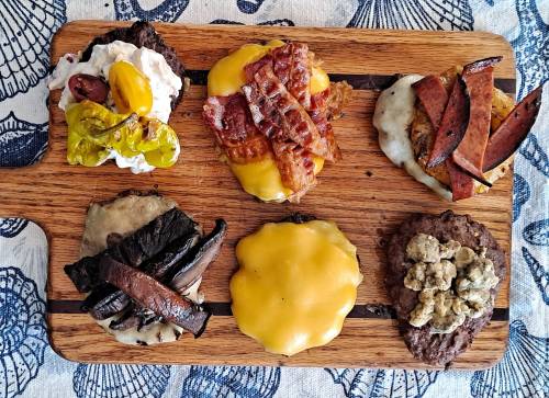 A wooden platter with various gourmet burgers topped with cheese, bacon, sausage, and unique condiments.