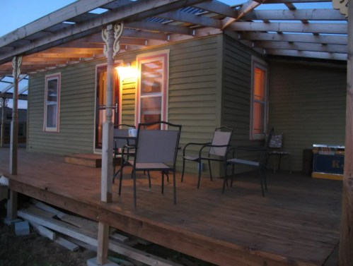 A cozy green house with a wooden porch, featuring chairs and warm lighting at dusk.