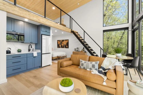 Modern living space with a cozy sofa, kitchen, and large windows overlooking greenery. Stylish staircase leads to a loft.
