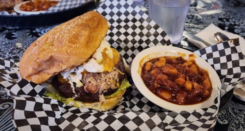 A hearty burger with pulled pork, lettuce, and a fried egg, served with a side of baked beans in a bowl.