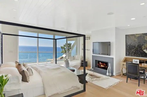 Modern bedroom with ocean views, featuring a large bed, fireplace, and stylish decor. Bright and airy atmosphere.