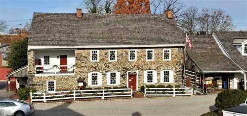 A historic stone house with a sloped roof, white trim, and red doors, surrounded by greenery and a cobblestone path.