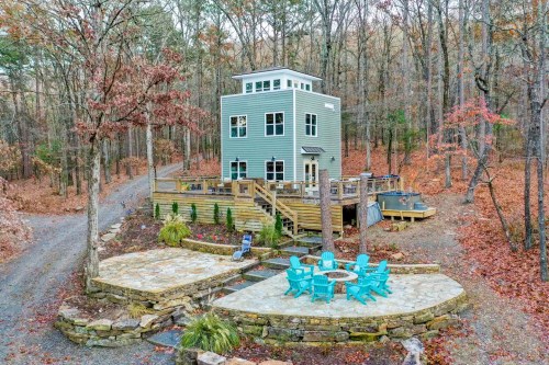 A modern, multi-story house surrounded by trees, with a stone patio and blue chairs in the foreground.