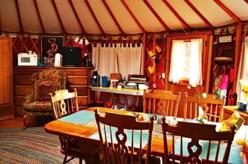 Cozy interior of a yurt featuring a dining table, chairs, kitchenette, and warm wooden decor.