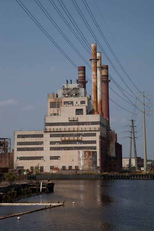 An industrial building with smokestacks beside a body of water, surrounded by power lines and a clear blue sky.