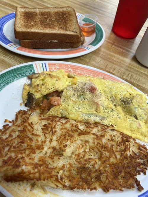 A plate with an omelet filled with vegetables, crispy hash browns, and a side of toast with a small cup of jelly.