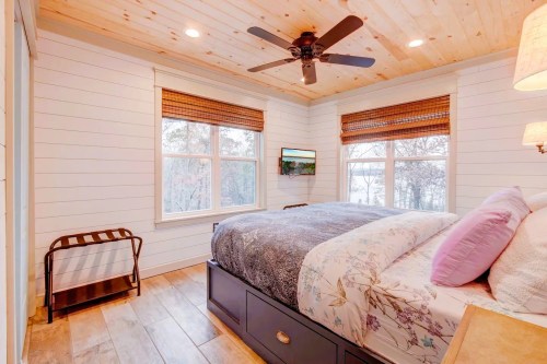 Cozy bedroom with a wooden ceiling, large windows, a bed with floral bedding, and a fan overhead.