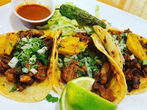 Three tacos filled with meat, onions, and cilantro, served with lime, lettuce, a jalapeño, and a side of salsa.