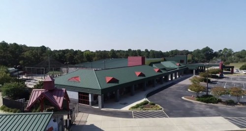 Aerial view of a large building with a green roof, surrounded by a parking lot and trees in the background.