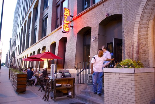 A bustling restaurant exterior with outdoor seating, red umbrellas, and a neon sign, as patrons enjoy the evening.