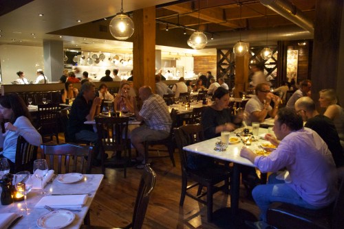 A bustling restaurant interior with diners at tables, warm lighting, and an open kitchen in the background.
