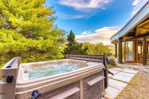 A hot tub on a stone patio surrounded by trees, with a scenic view and a modern building nearby under a blue sky.