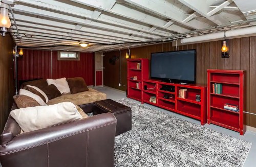 Cozy basement living area with a brown couch, red bookshelves, a large TV, and a patterned rug.