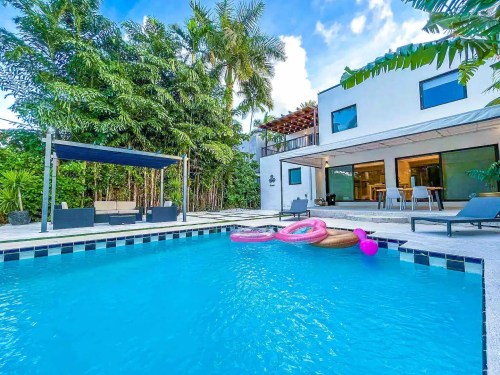 A bright pool area with pink floaties, surrounded by lush greenery and a modern house in the background.