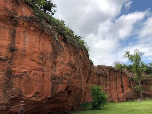 A large red rock formation with greenery at the base and a cloudy blue sky above.