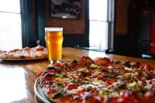 A close-up of a pizza on a wooden table, with a pint of beer in the background and another pizza in the distance.