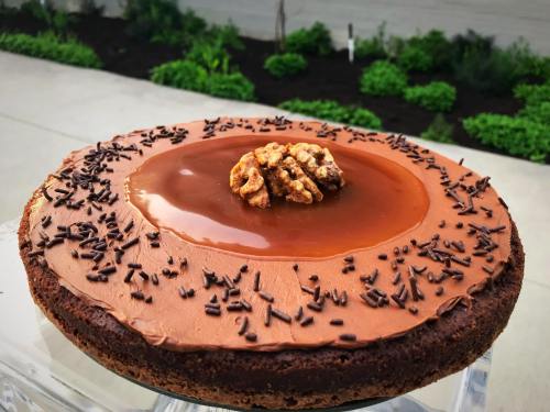 A chocolate cake topped with caramel, walnuts, and chocolate sprinkles, set against a green garden backdrop.