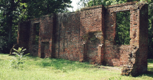 Ruins of a brick building overgrown with vegetation, surrounded by trees and grass.