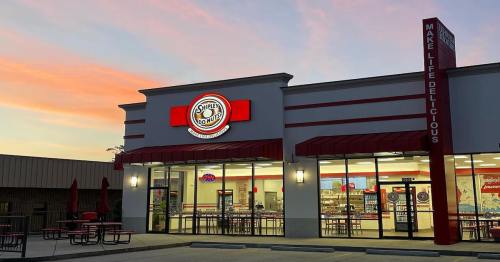A brightly lit donut shop exterior at sunset, featuring a red and white sign and outdoor seating.