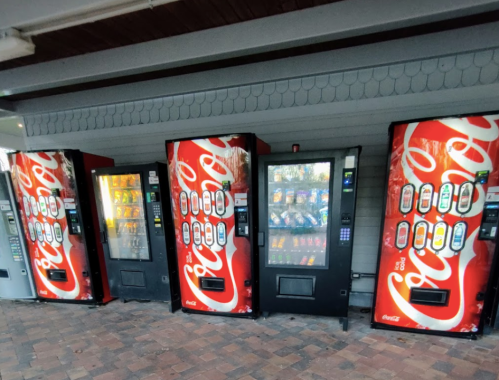 A row of vending machines, including snack and beverage options, against a gray wall.