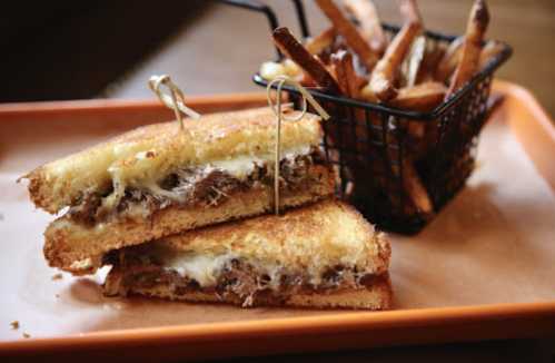 A grilled cheese sandwich with pulled meat, served with a side of crispy French fries in a small basket.